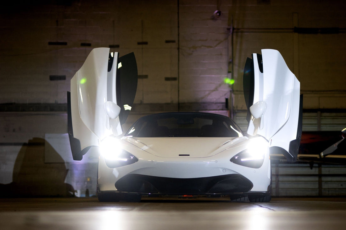 A white McLaren spreads its dihedral doors under purple ambient lighting inside the hangar.