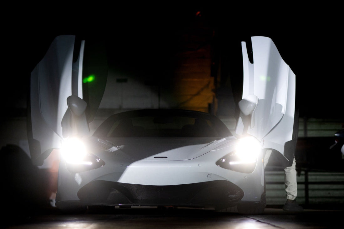 A white McLaren spreads both dihedral doors wide open in a dark hangar with its headlights blazing.