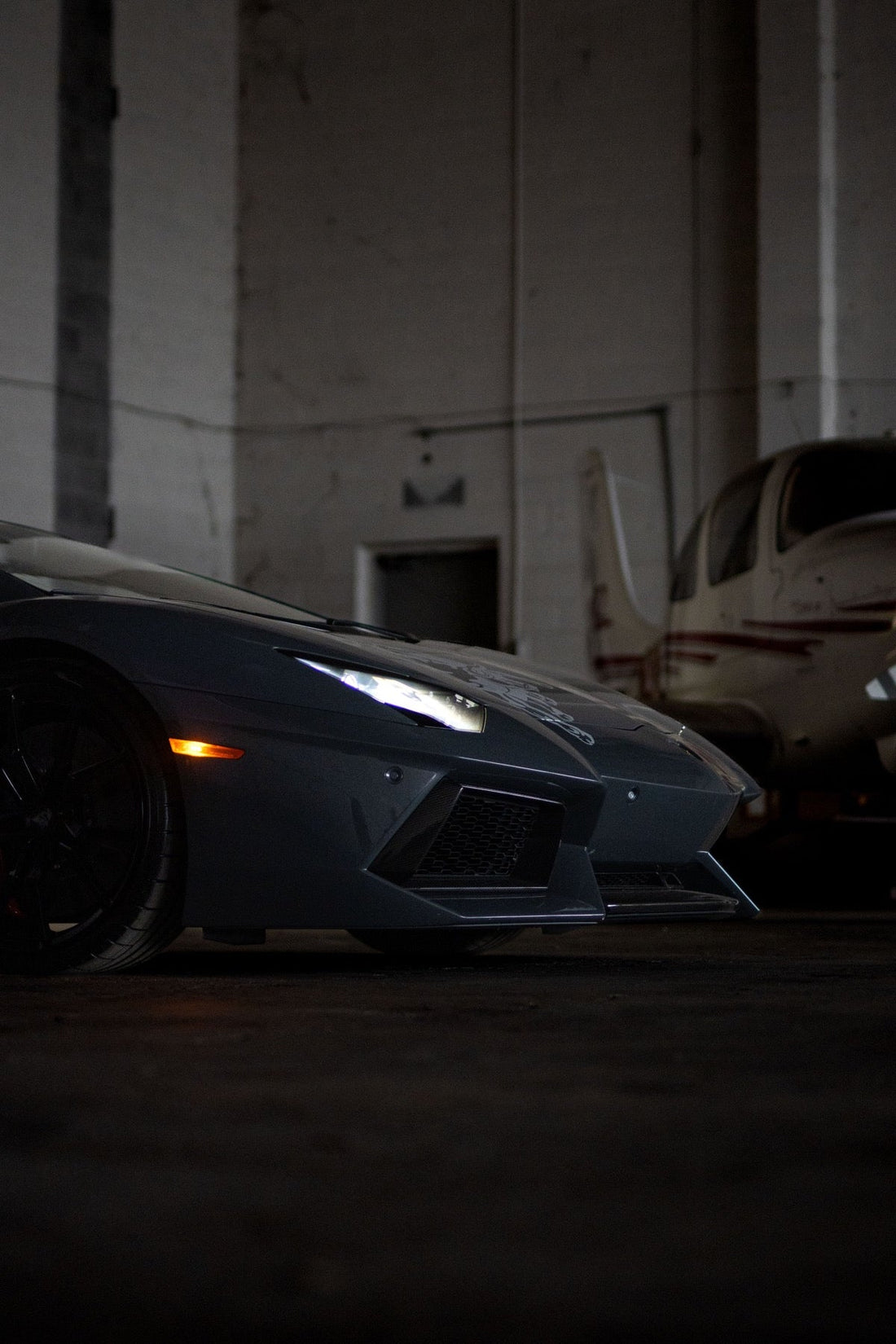A grey Lamborghini Aventador is parked nose to nose with a small propeller plane inside a hangar.