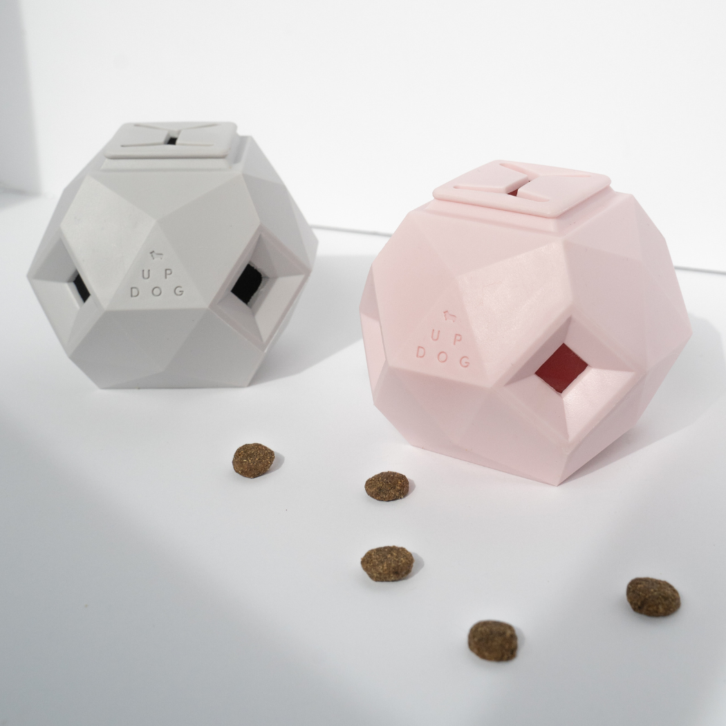 Two Odin treat dispensing dog toys by Up Dog Shop, one light grey and one pink, faceted with open treat windows and treats scattered nearby