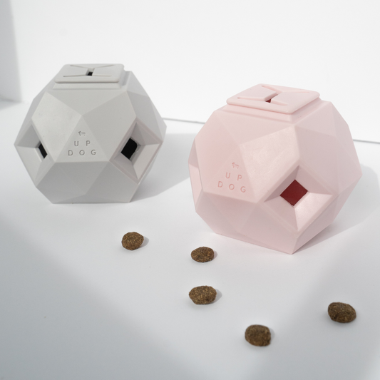 Two Odin treat dispensing dog toys by Up Dog Shop, one light grey and one pink, faceted with open treat windows and treats scattered nearby