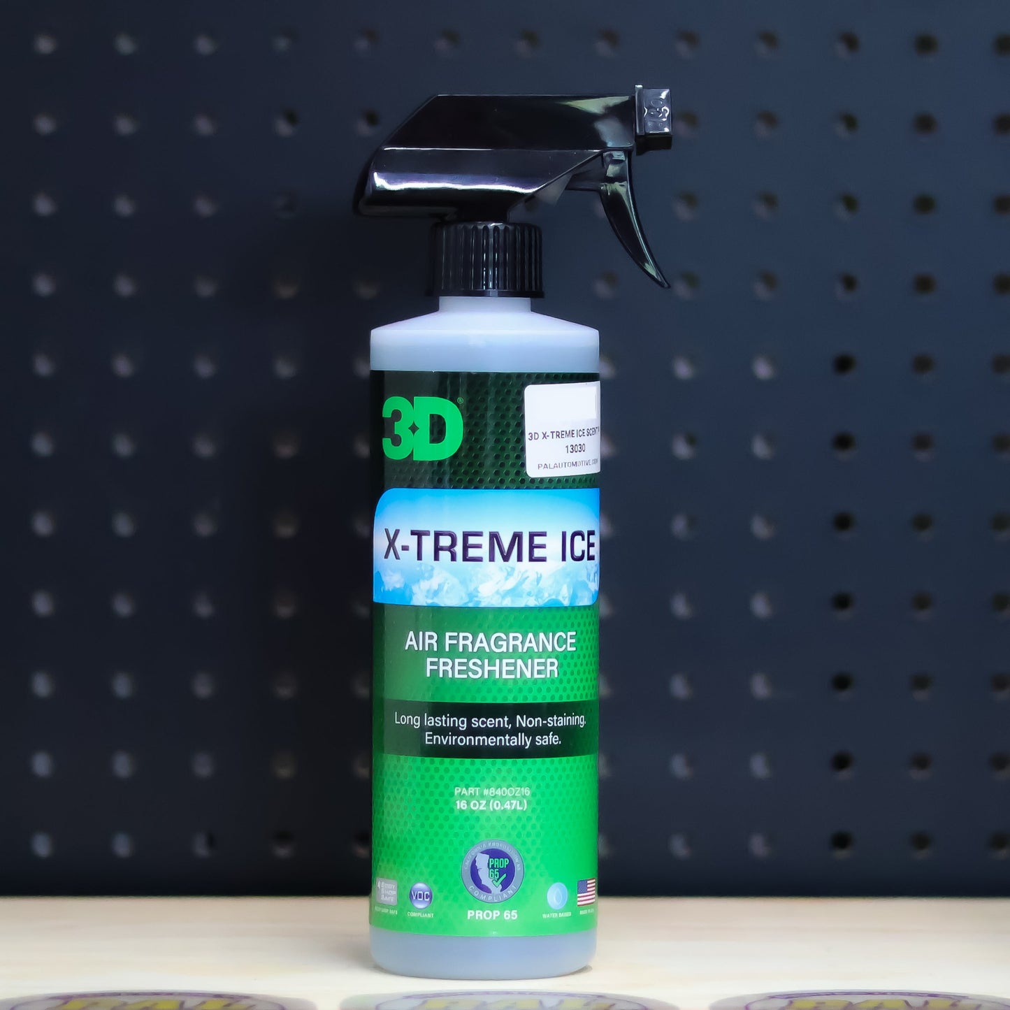 3D X-Treme Ice air freshener pint bottle, blue and green label with a black trigger sprayer, on a bench in front of a dark pegboard